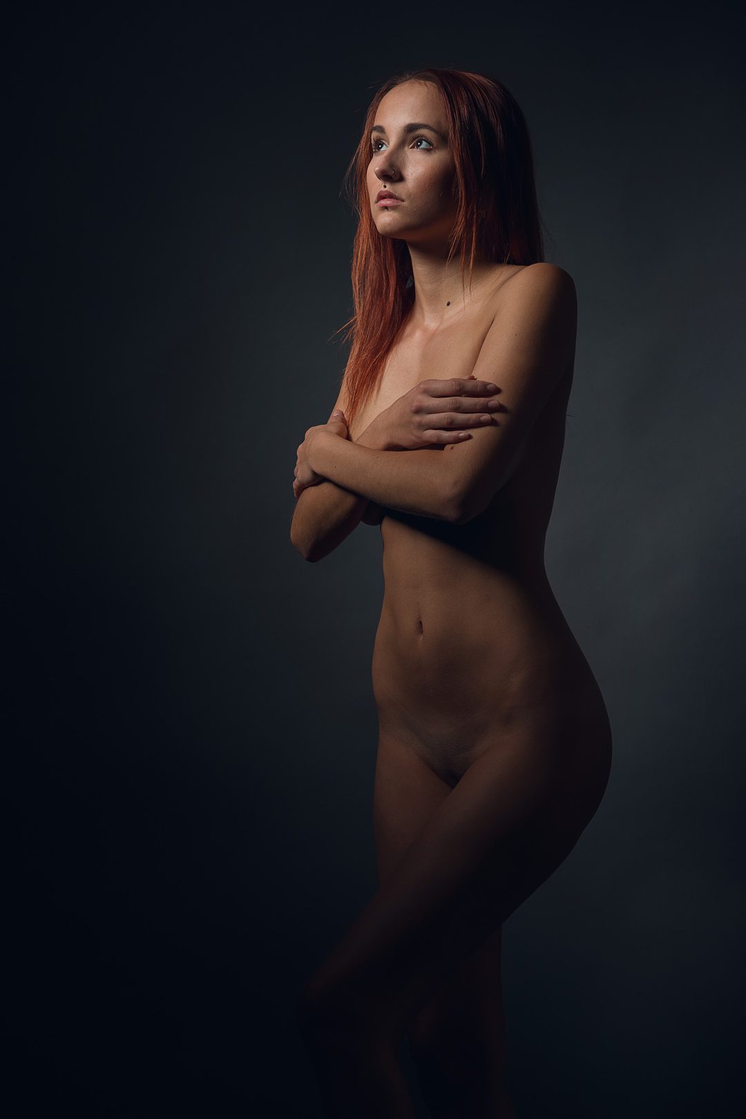 Künstlerischer Studioakt der Modelin Lucia, stehend mit verschränkten Armen vor dunklem Hintergrund, Bratislava, Slowakei, 2018.